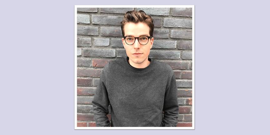 Tobias Rydén Sjöstrand, ung man med kort mörkt hår, glasögon och grå tröja, som står lutad mot en tegelvägg.