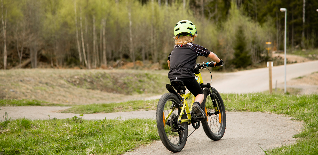 En pojke cyklar på en mountainbikecykel i ett pumptrackspår på Borgen Outdoor. Gräset och träden är gröna.