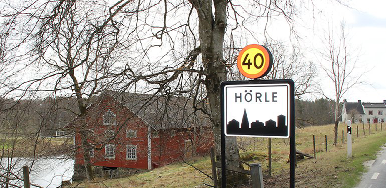 Ett rött hus står vid en sjö och en väg. Framför huset står en skylt med texten Hörle.