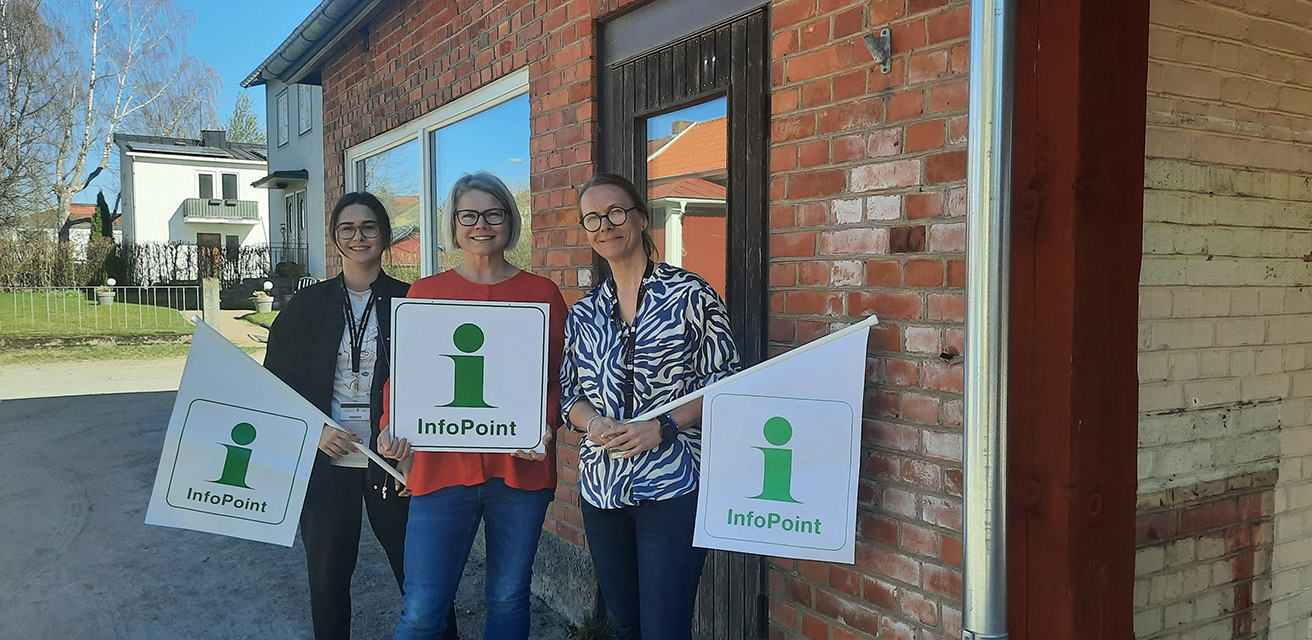 Tre kvinnor står framför ett litet hus i tegel med stora fönster. Kvinnorna håller i flaggor med texten "InfoPoint", och med ett grönt stort I på en vit bakgrund.