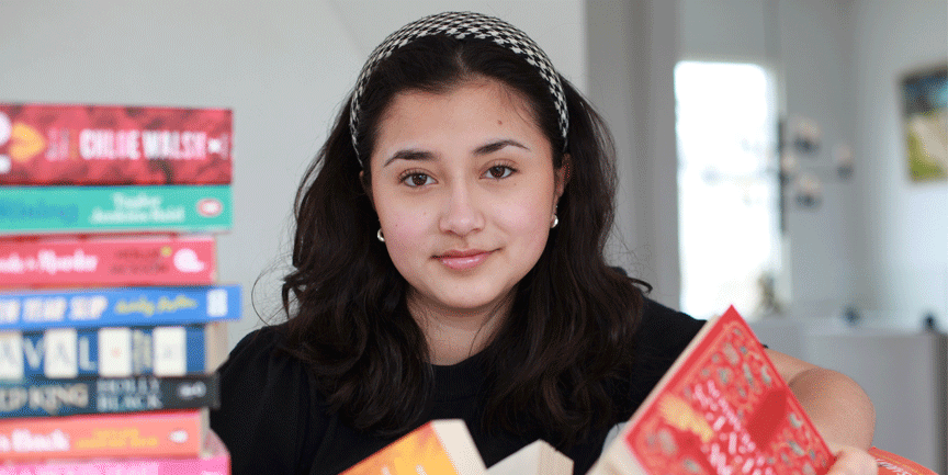 Isabella, Bellalovesbooks, ung kvinna med långt mörkt hår som är omgiven av böcker.