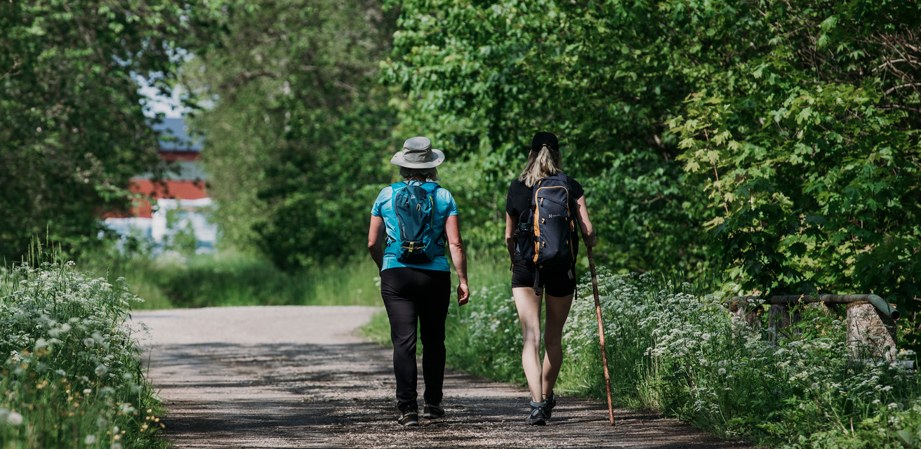 Två kvinnor vandrar en sommardag och går med hatt och vandringskäpp.
