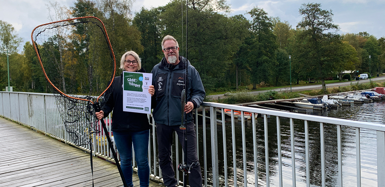 Karin Fritz, Värnamo kommun och Hans Sågman, Sons of Anglers, står på bron på Osudden och håller i en håv, ett fiskespö, och en affisch för evenemanget Gäddhänget.