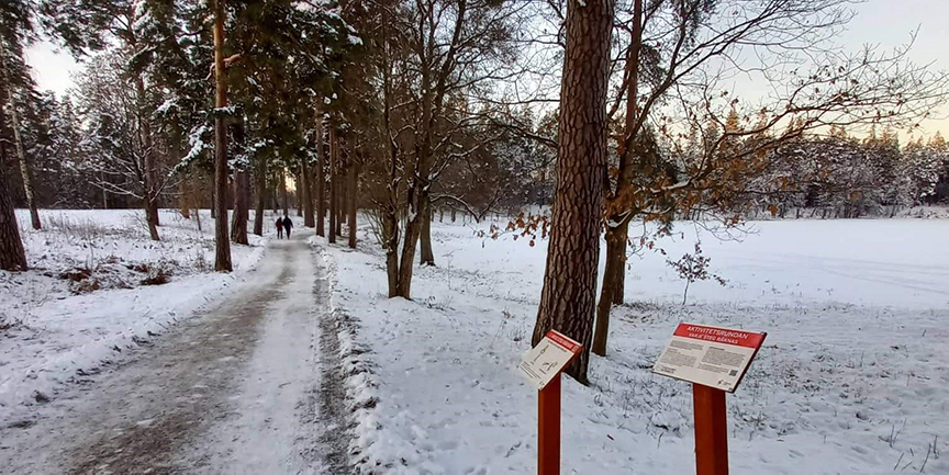 En motionsrunda vintertid. Snö på backen och träden. I förgrunden ses skyltar för hälsorundan. I bakgrunden syns två personer som går på promenad.