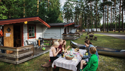 En familj sitter runt ett bord utomhus och äter mat på campingen.