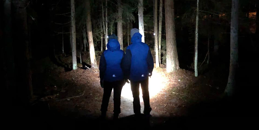 Två personer står i mörkret och lyser med ficklampa mot skogen.
