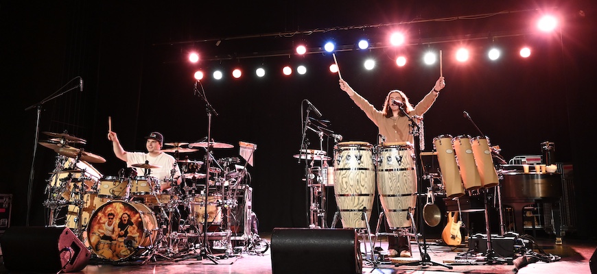 Två personer på en scen med spotlights. En person till vänster spelar trummor och sträcker upp händerna i luften. En person till höger spelar percussion och sträcker upp händerna i luften. Både spelar på 3d-printade trummor.