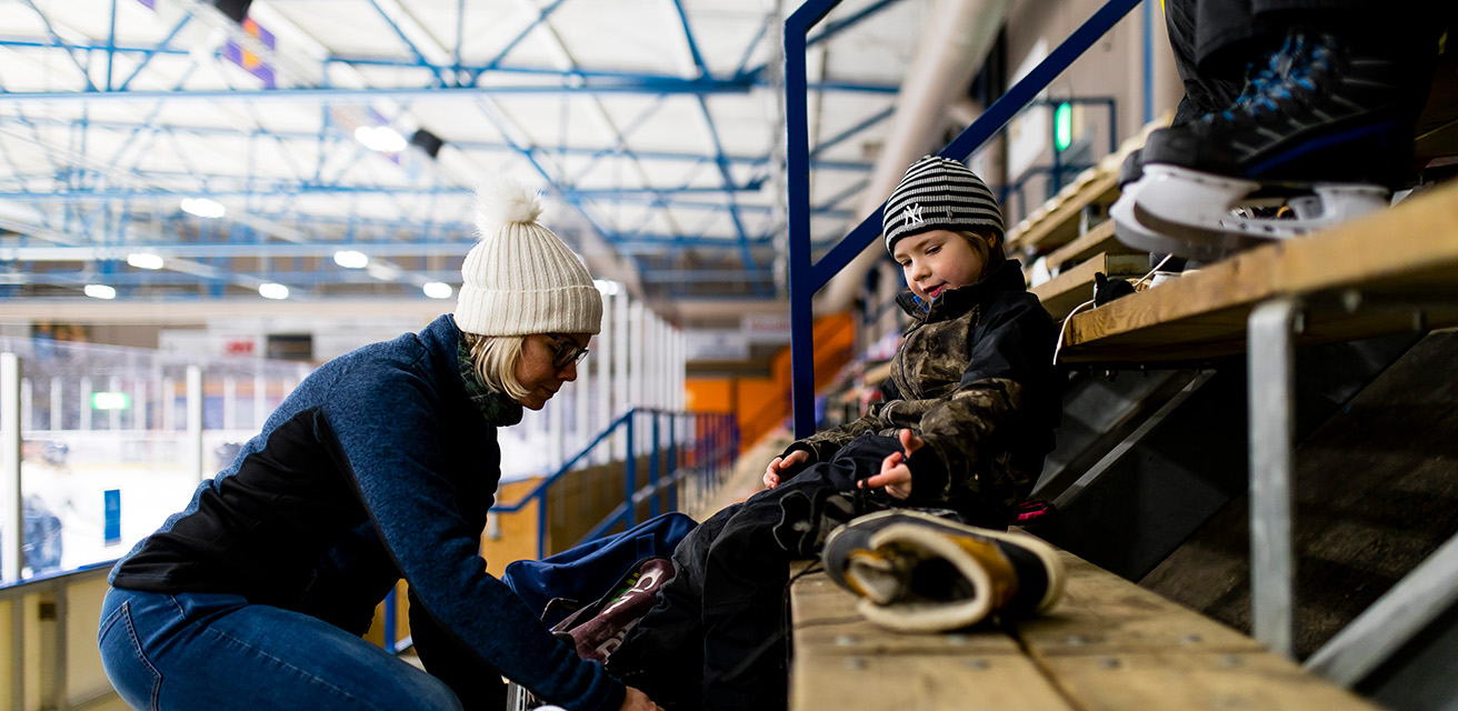 En kvinna knyter ett barns skridskor. De sitter på en läktare vid en ishall.