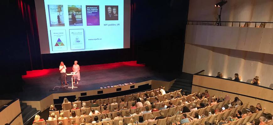 Vy över scenen på auditoriet i Gummifabriken där två kvinnor föreläser och det är fullt i bänkraderna med publik.