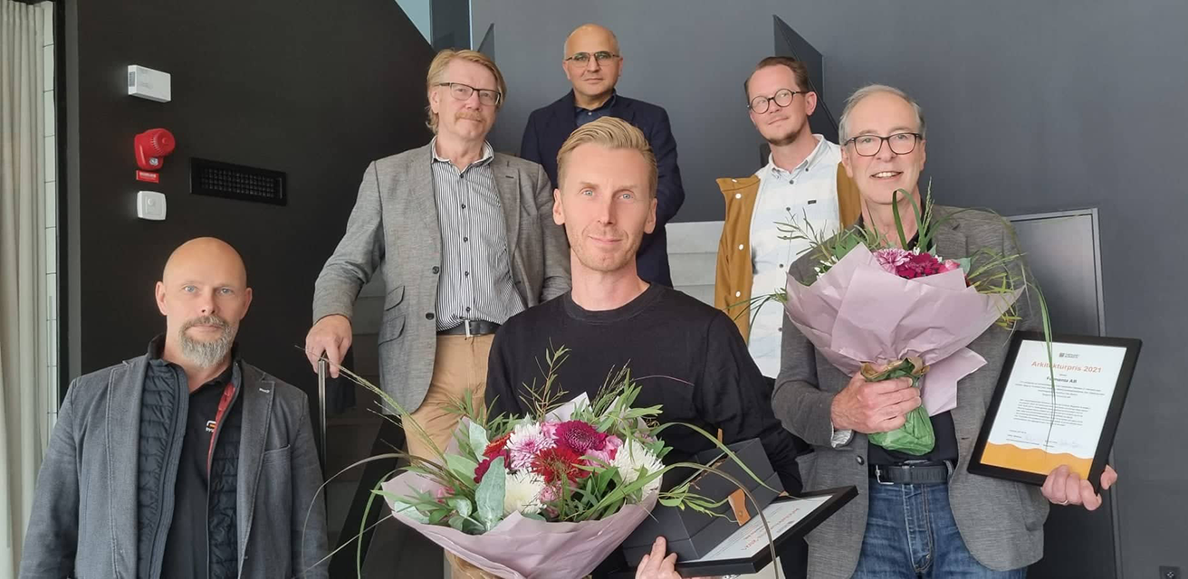 Sex män från Formenta som tar emot kommunens arkitekturpris 2021. De står inomhus och ser glada ut, och bär på ett diplom och två buketter.
