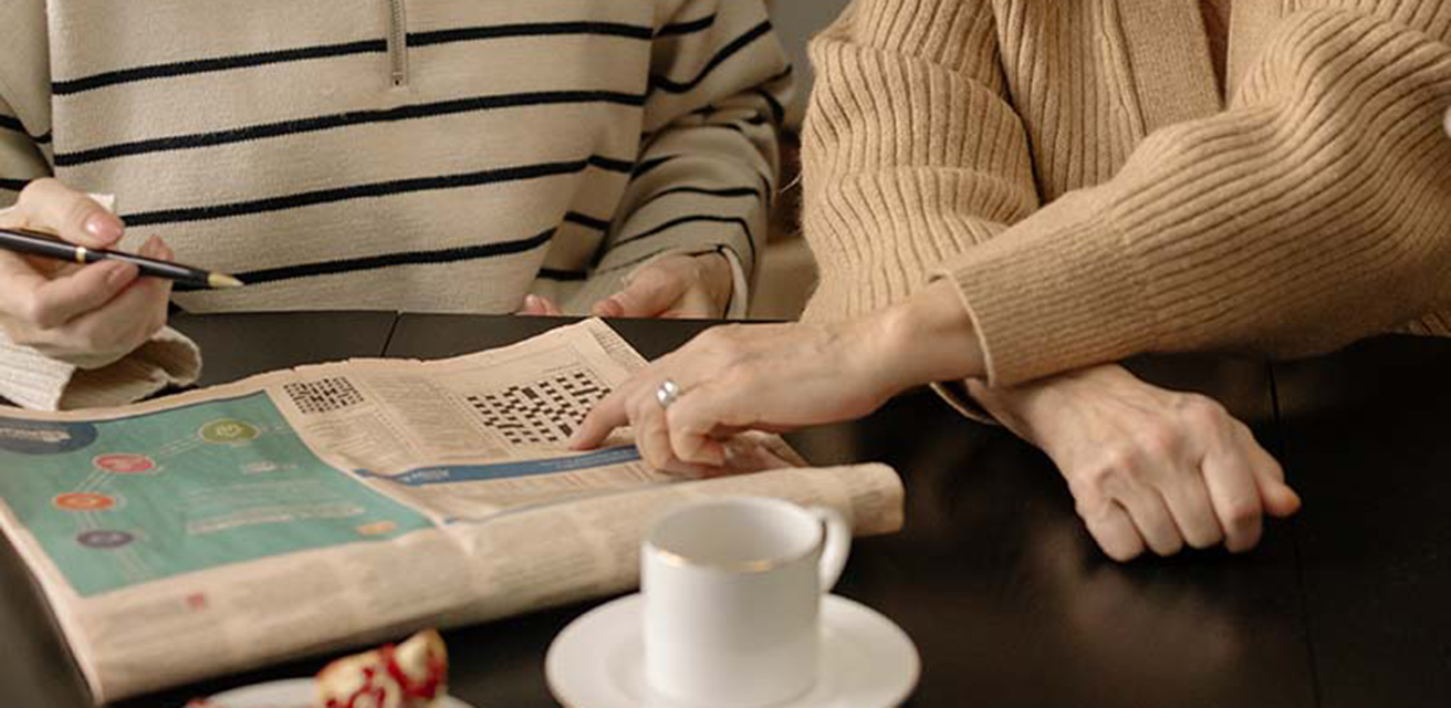 Två personer sitter vid ett bord. På bordet finns en tidning, en bulle och en kopp. De löser korsordet i tidningen tillsammans.