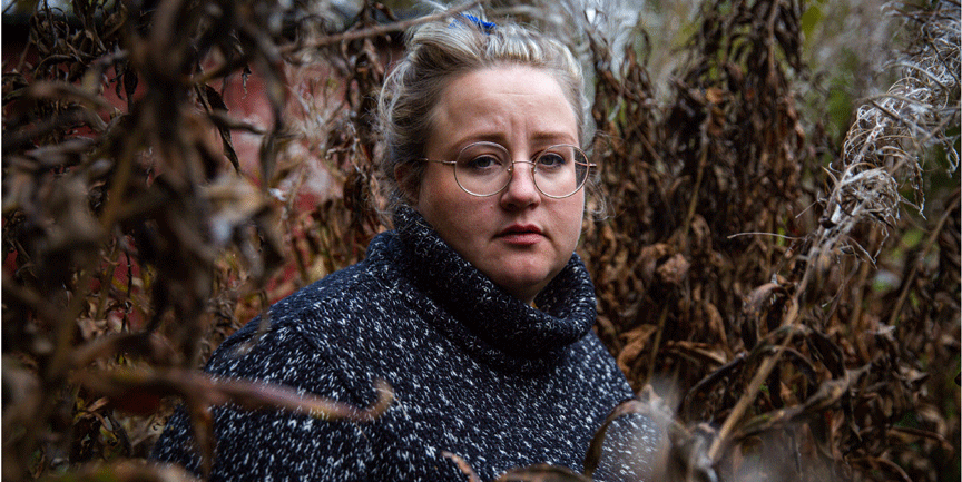 Ellen Strömberg, kvinna med glasögon och blå stickad sjal, som står bland höstiga bruna buskar.