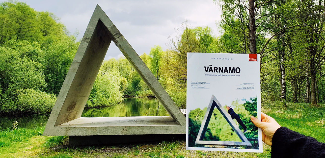 En hand håller i Värnamo Upplevelseguide. På omslaget finns en bild på bänken Polygon. Bakom guiden ser man bänken Polygon.