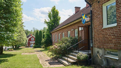Bilden föreställer en röd tegelbyggnad med blommande grön trädgård