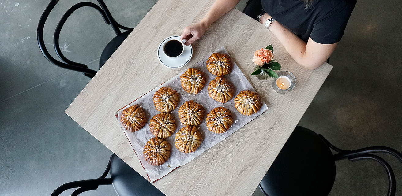 Bild tagen ovanifrån på ett bord med en plåt kanelbullar, en blomma och en kaffekopp. En kvinna sitter och håller i koppen.
