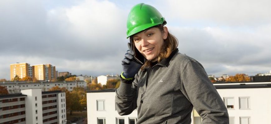 Kvinna med bygghjälm står på ett hustak och pratar i mobiltelefon. I bakgrunden vy över tak på flera flerfamiljshus.